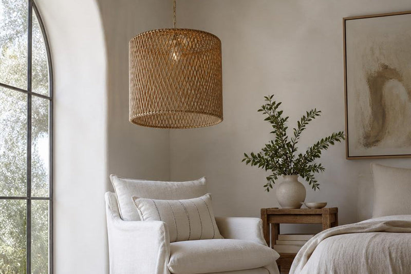 A Drum Rattan Pendant Light hanging in a calm bedroom corner above a white linen reading chair, with a made bed and framed art nearby