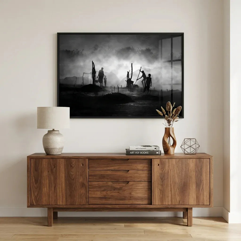Black-and-white photographic print of standing figures in soft early-morning mist, in a black frame above a walnut sideboard styled with a ceramic lamp and dried grasses