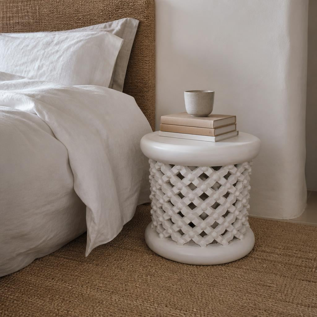 White Bamileke stool used as a bedside table beside a linen bed